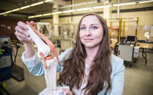 kobieta z długimi włosami prezentuje prototyp stawu kolanowego, w tle urządzenia w hali technologicznej