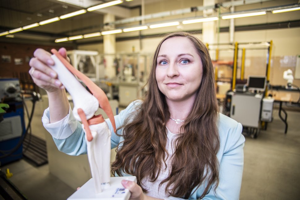 kobieta z długimi włosami prezentuje prototyp stawu kolanowego, w tle urządzenia w hali technologicznej
